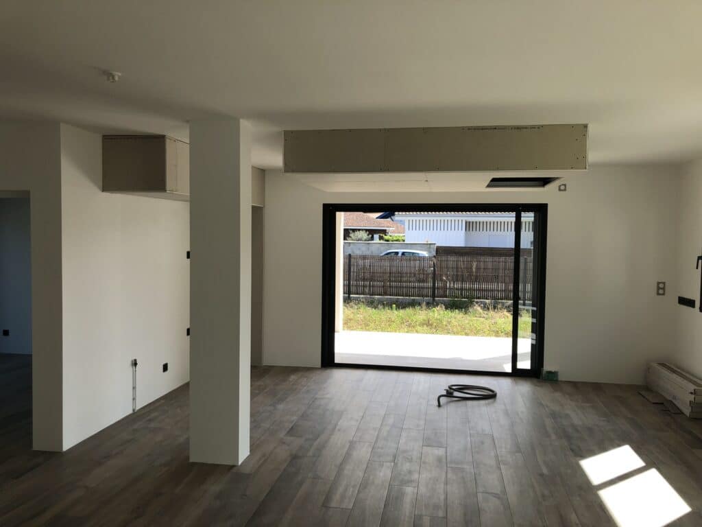Aménagement de salon moderne : baie vitrée et sol bois Intérieur de maison en travaux avec carrelage aspect bois, pilier blanc et grande baie vitrée ouvrant sur un jardin.